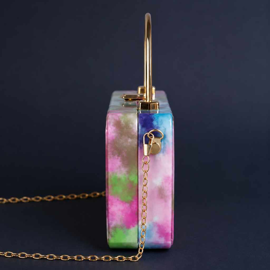 A square resin clutch bag with a colorful tie-dye pattern in pink, blue, green, and white, featuring a gold-tone top handle, lock, and chain strap, set against a dark navy background.