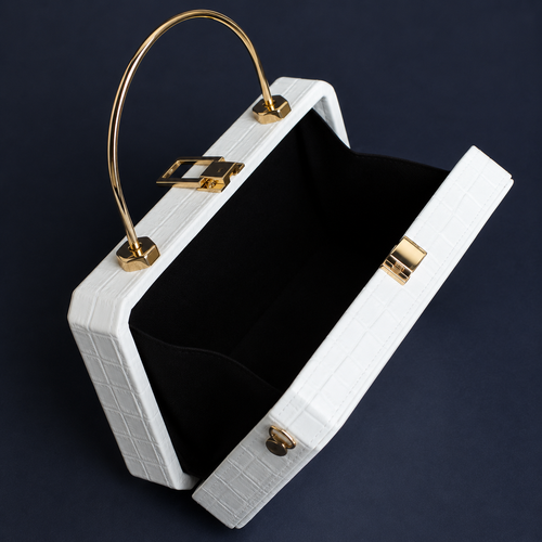 A structured white crocodile-embossed handbag with a black velvet interior and gold-tone hardware, shown open against a dark navy background.