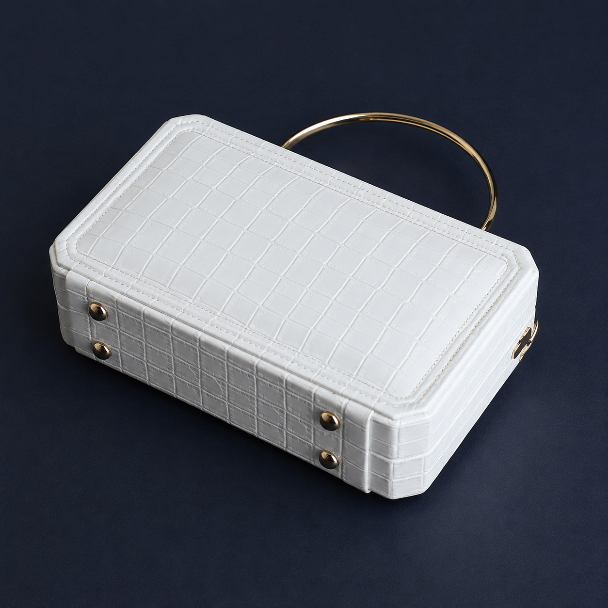 A white faux-crocodile leather handbag with a gold-tone metal handle and gold hardware, photographed on a dark navy background.