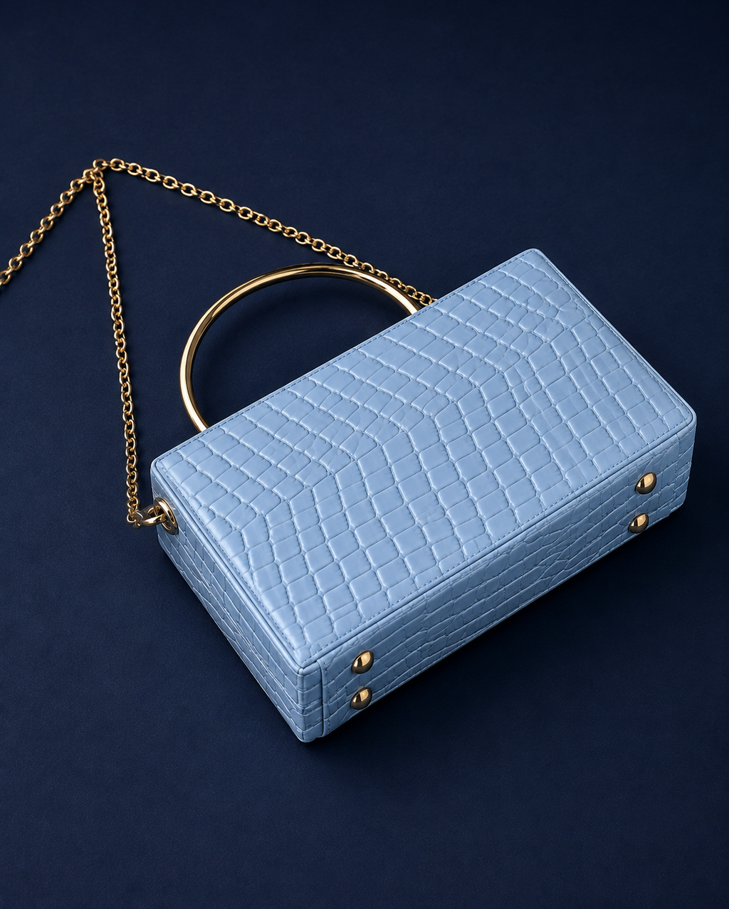 A light blue crocodile-embossed leather handbag with a gold-tone chain strap and a circular gold handle, set against a dark navy blue background.