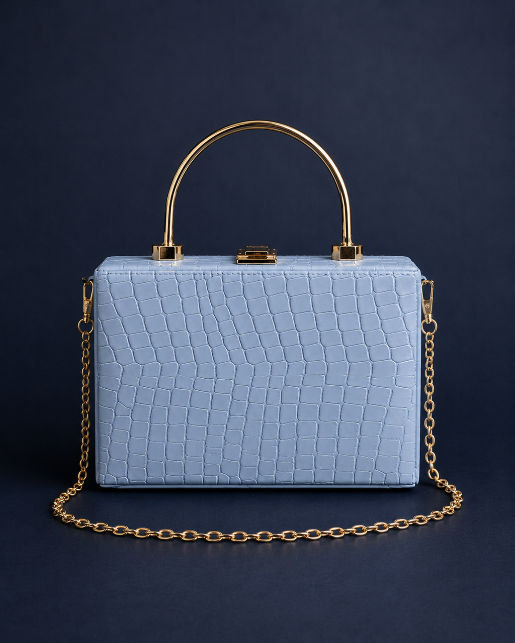 A light blue crocodile-embossed leather handbag with a gold-tone chain strap and a circular gold handle, set against a dark navy blue background.