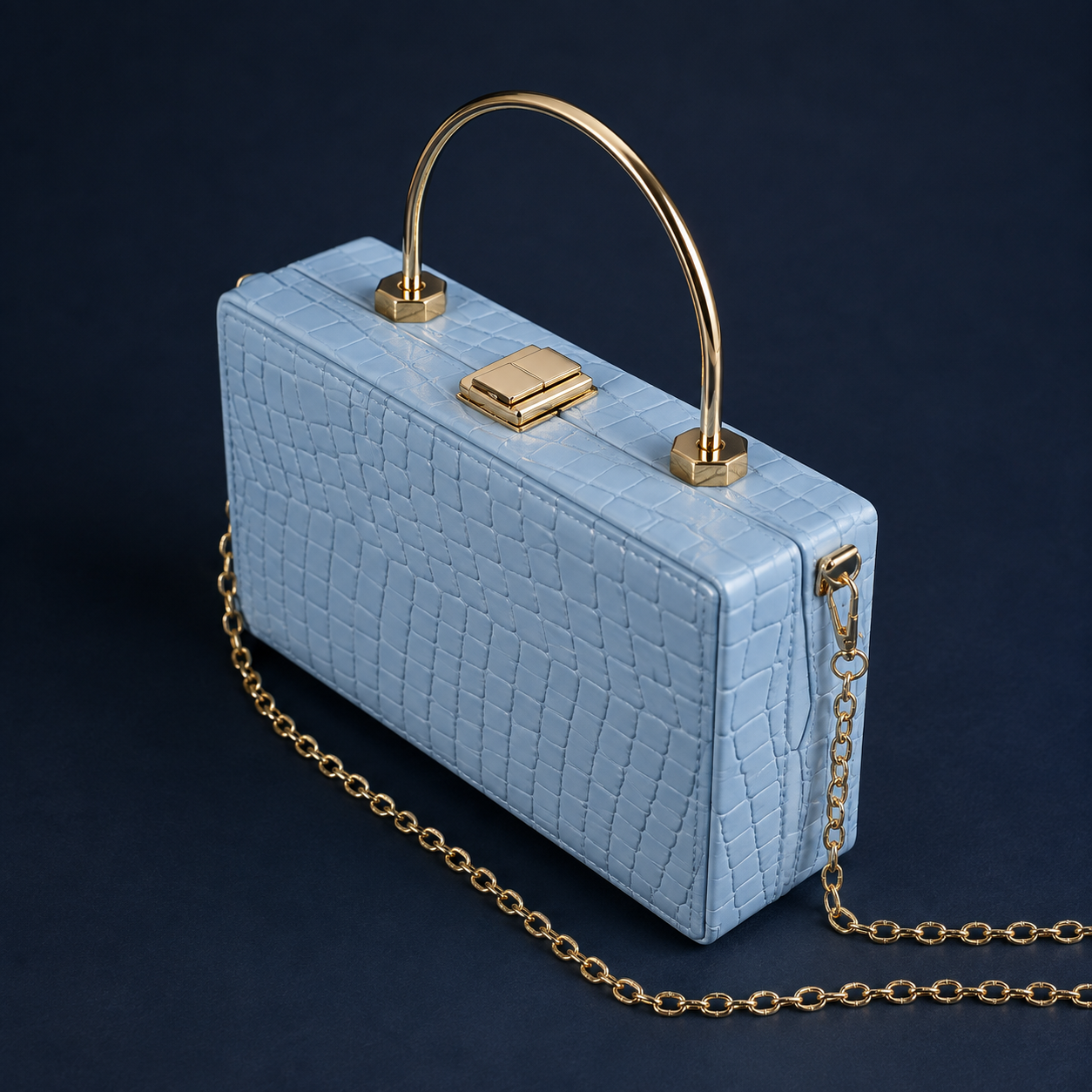A light blue crocodile-embossed clutch bag with a gold arched top handle and a gold chain strap, set against a dark navy background.