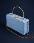 A light blue crocodile-embossed leather handbag with a gold-tone arched top handle and chain strap, set against a dark navy background.