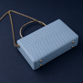 A light blue crocodile-embossed clutch bag with a gold-tone chain strap and a circular top handle, set against a dark navy background.