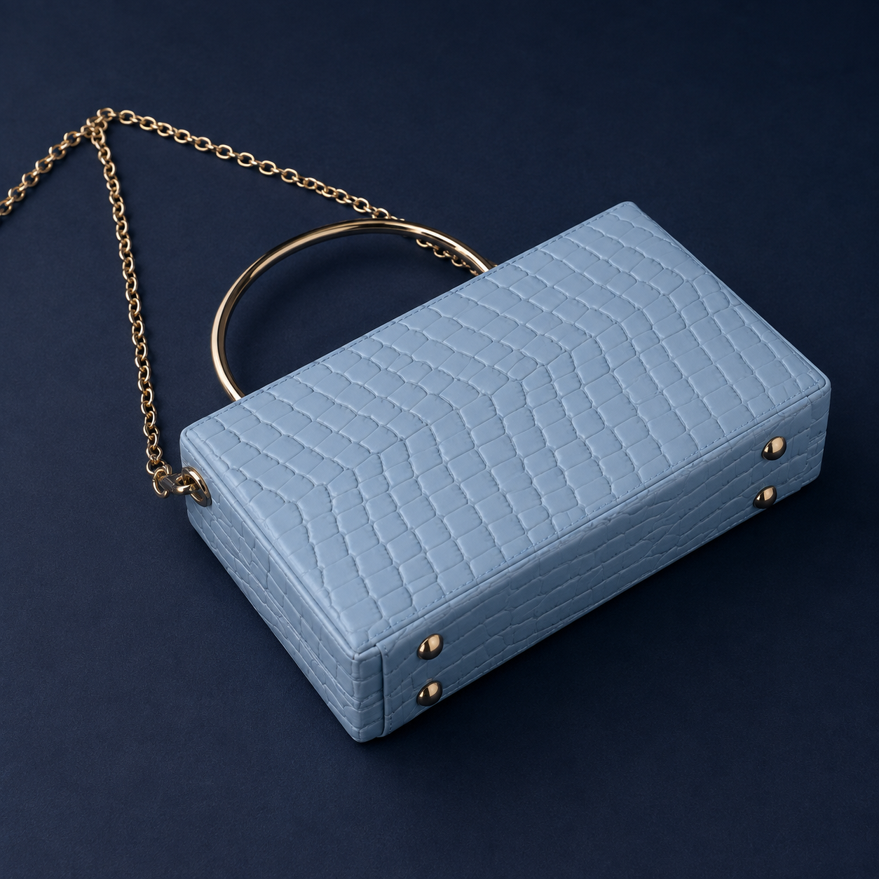 A light blue crocodile-embossed clutch bag with a gold-tone chain strap and a circular top handle, set against a dark navy background.