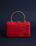 A red textured clutch bag with gold metallic veining and a curved gold metal top handle, set against a dark navy blue background.
